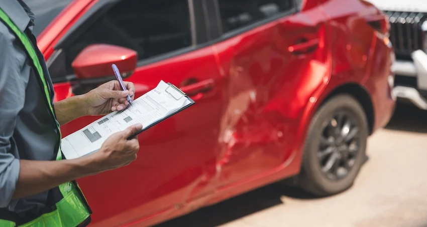 An insurance adjuster in a green safety vest documents damage on a clipboard beside a red sedan with a heavily dented door- representing the on-site vehicle inspection that kicks off the car accident claim process- with help from Panchenko Law Firm- after a collision in Charlotte NC.