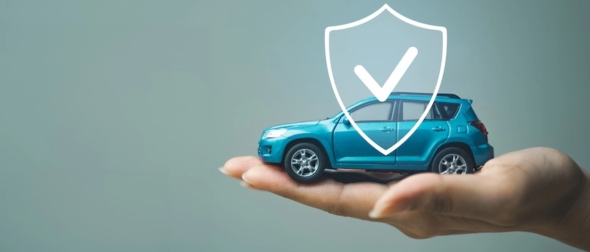 A hand holds a small blue SUV toy with a white shield and checkmark graphic overlaid on top against a gray background- symbolizing the insurance protection that supports drivers throughout the car accident claim process after an unexpected collision in Spartanburg SC.