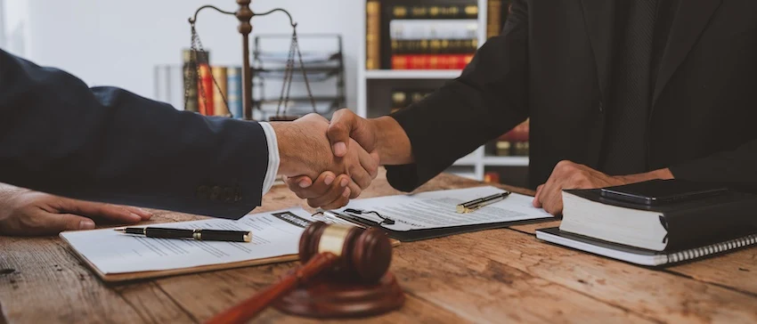 An attorney from Panchenko Law Firm shakes hands with a client across a rustic wooden desk covered with signed legal documents- a gavel- and a pair of scales of justice visible on the bookshelf behind them- representing the agreement reached when an injury victim hires a car accident lawyer to pursue their case.