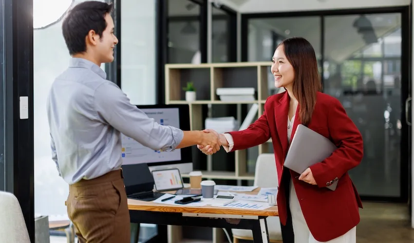 A smiling female attorney from Panchenko Law Firm in a red blazer holds a laptop and shakes hands with a male client in a modern office- capturing the positive outcome an accident victim experiences when their car accident lawyer successfully negotiates maximum compensation on their behalf in Spartanburg SC.