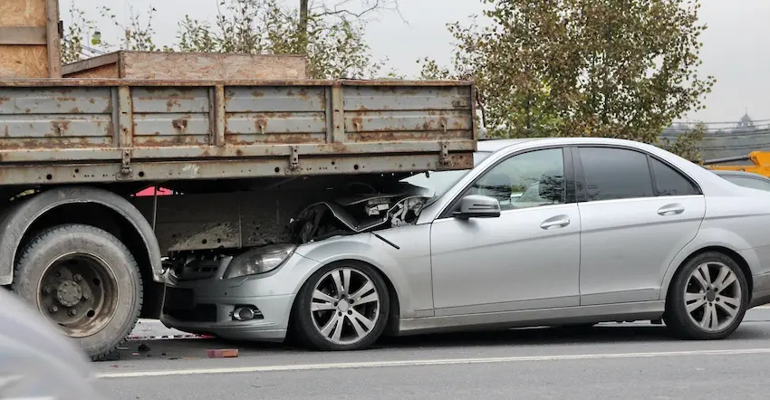 A silver sedan with a severely crushed front end is lodged underneath the rusted flatbed of a large commercial truck on a city road representing the type of catastrophic vehicle underride collision that results in serious car accident claims against commercial trucking companies in Charlotte NC.