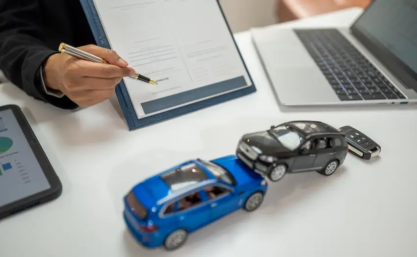 A person in a dark blazer points a pen to a document on a clipboard beside two model cars staged in a rear-end collision-a set of car keys-a tablet-and a laptop on a white desk representing the paperwork and documentation involved in filing car accident claims with an attorney from Panchenko Law Firm or an insurance company in Spartanburg SC.