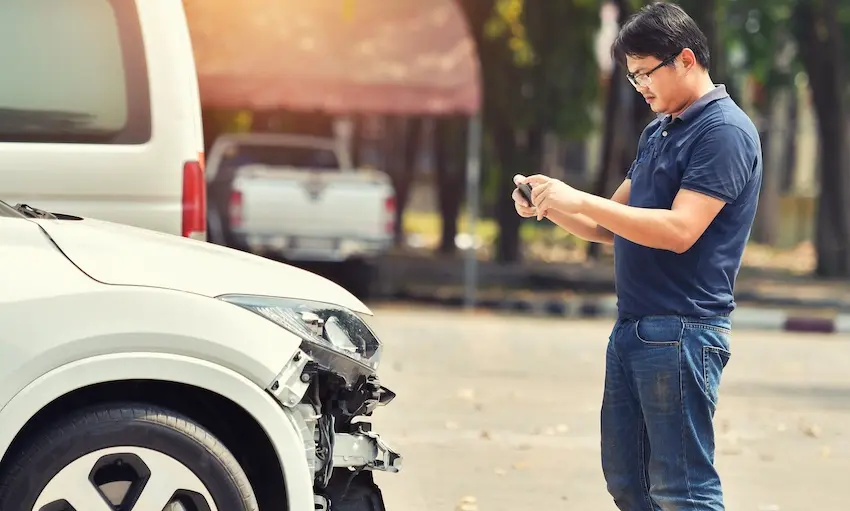 A man in a navy polo shirt photographs the crumpled front end of a white car with his smartphone on a roadside capturing the critical evidence-gathering step that supports a successful car accident insurance claim after a collision with help from Panchenko Law Firm.