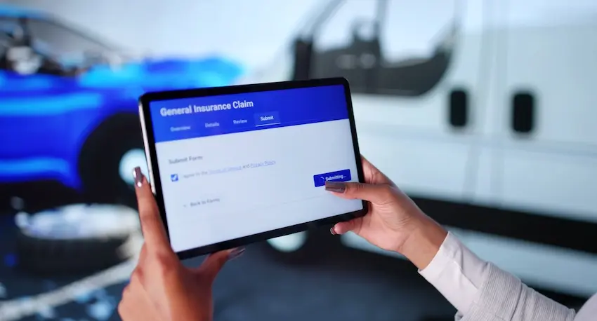 A woman holds a tablet showing a "General Insurance Claim" submission form in the active process of being submitted with vehicles blurred in the background illustrating the digital filing step that initiates a car accident insurance claim after a crash in Spartanburg SC.