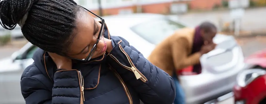 A woman with long braids and glasses stands in the foreground with her head bowed and hand pressed to her neck in pain while another person in a brown jacket sits distressed in the background beside damaged vehicles on an overcast street depicting two accident victims in the immediate aftermath of a collision who may be entitled to compensation benefits for their injuries and losses in Charlotte NC.