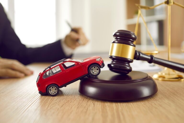 A bright red toy SUV is propped at a dramatic angle with its front end lifted off the surface and its rear wheels on the ground leaning against the circular dark mahogany sound block of a judge's gavel with a wide gold band that stands upright beside it on a warm wooden desk- while a blurred professional in a dark suit sits in the background holding a pen over documents with one hand resting on the desk and an ornate brass balance scale of justice is partially visible on the right side of the frame in soft focus — a richly detailed legal concept image representing the courtroom advocacy a car accident attorney provides to injured clients seeking fair compensation in Charlotte NC.