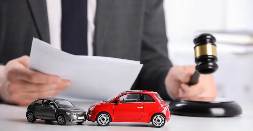 A gray toy sedan and a red compact toy car are positioned nose-to-nose in a head-on collision on a white desk surface in the sharp foreground while a professional in a dark gray suit sits slightly blurred directly behind them- holding a white legal document open in one hand and gripping a dark wooden judge's gavel with a gold band in the other hand poised above its sound block — a composed legal concept image representing the active role a car accident attorney from Panchenko Law Firm in Charlotte NC plays in reviewing case documents and pursuing justice on behalf of clients injured in a collision.